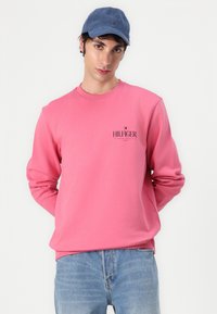 Rosa genser med lange ermer, rund hals; har brodert "HILFIGER"-logo og liten logo-detajle. Brukt med lyseblå jeans og en blå caps.