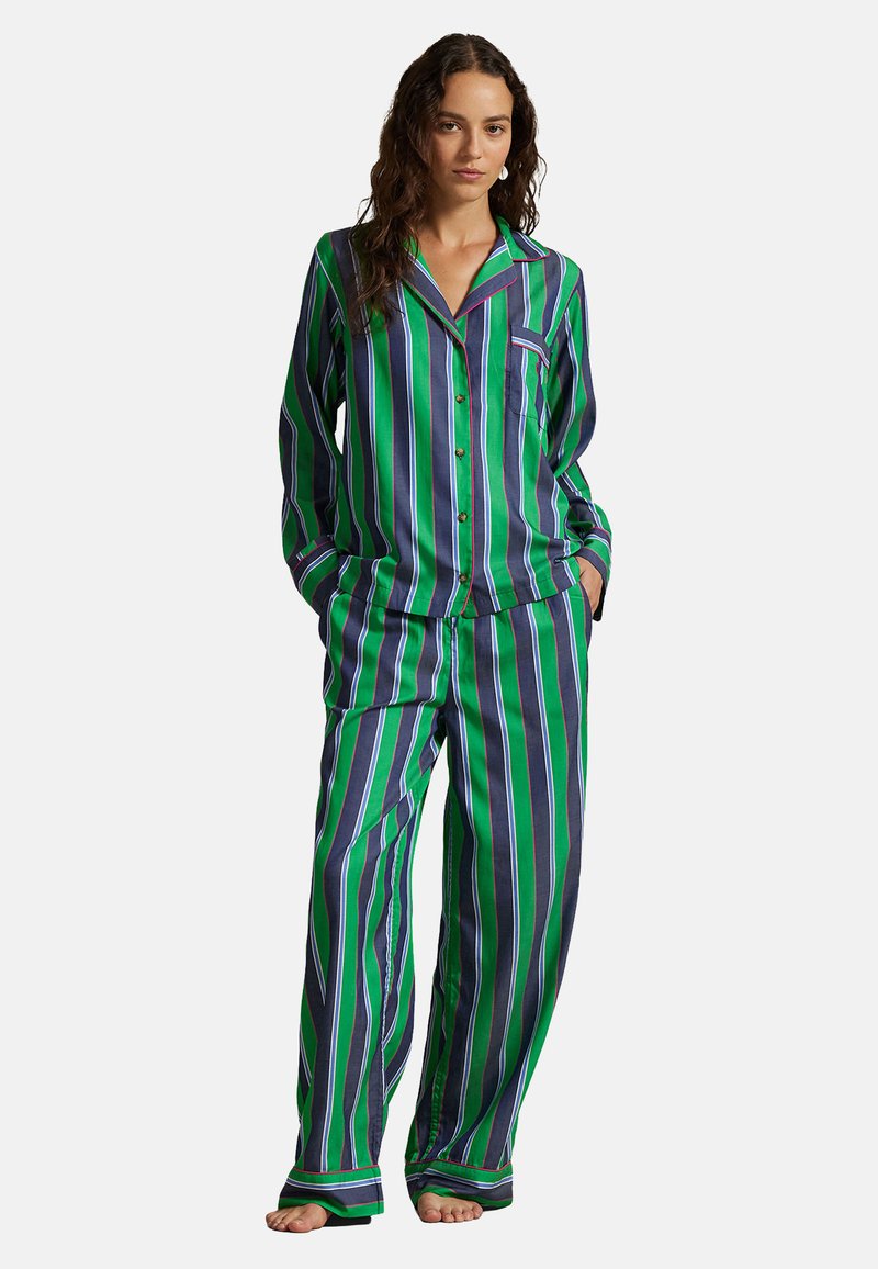 Frau steht barfuß und trägt ein Pyjama-Set aus grün, dunkelblau und lila vertikal gestreiften, langärmligen Hemd und Hose.