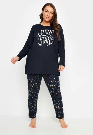 Marineblå langærmet pyjamashirt med teksten "SKIN som STJERNEERNE", parret med matchende bukser med flerfarvede stjerne-mønstre.