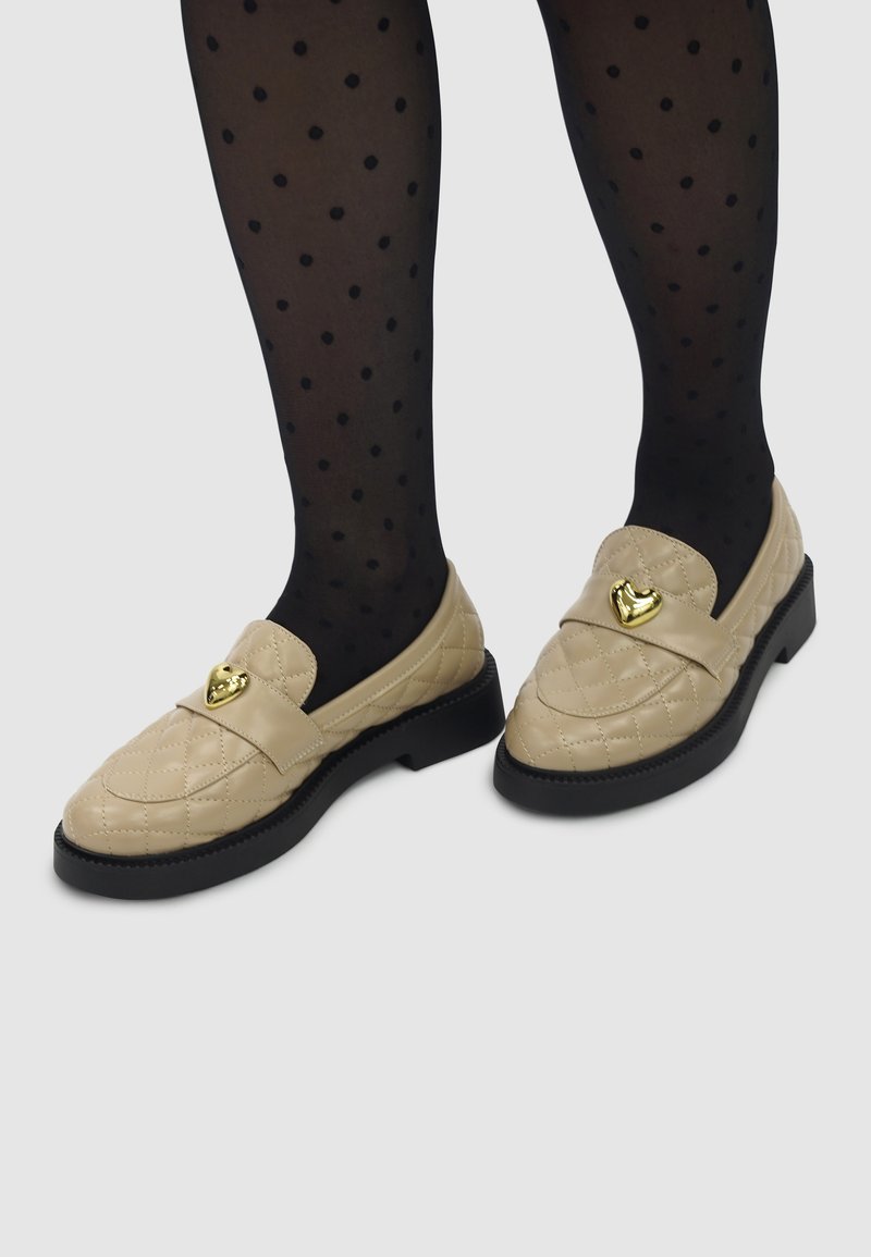 Beige gewatteerde loafers hebben een platte zwarte zool en een gouden hartvormige accent aan de voorkant. Draagt met zwart met stippen panty's.