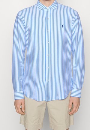 Uomo che indossa una camicia a righe verticali blu e bianca con maniche lunghe e pantaloncini beige, in piedi davanti a uno sfondo neutro.