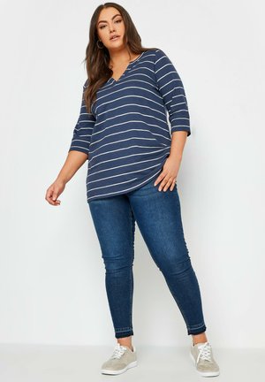 Gestreiftes navyblaues Oberteil mit V-Ausschnitt und ¾-Ärmeln, kombiniert mit blauen Skinny-Jeans und hellgrauen Sneakern. Weicher Stoff mit lockerer Passform.