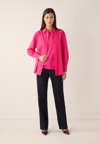 Fuchsiafarbene Bluse mit Knopfleiste und Tasche über einem passenden Oberteil, kombiniert mit dunklen Jeans und spitzen Absatzschuhen, vor neutralem Hintergrund.