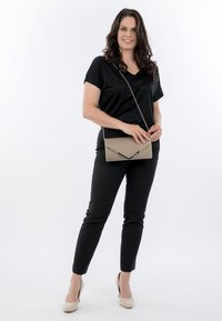 Mujer con top y pantalones negros, sosteniendo un bolso clutch beige con correa de cadena, de pie sobre fondo blanco, sonriendo.
