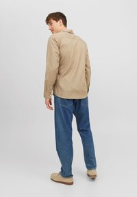 Beige long-sleeved shirt with a collared neckline and button cuffs, paired with loose blue jeans and light-colored footwear.