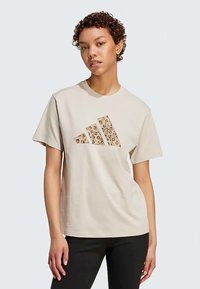 Ženska s kratkimi kodrastimi lasmi, oblečena v bež majico z leopardjim potiskom logotipa Adidas in črnimi hlačami pred nevtralno ozadjem.