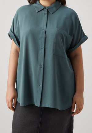 Femme portant une chemise ample turquoise à manches courtes, boutonnée, avec une poche poitrine et des manches retroussées, associée à une jupe sombre.
