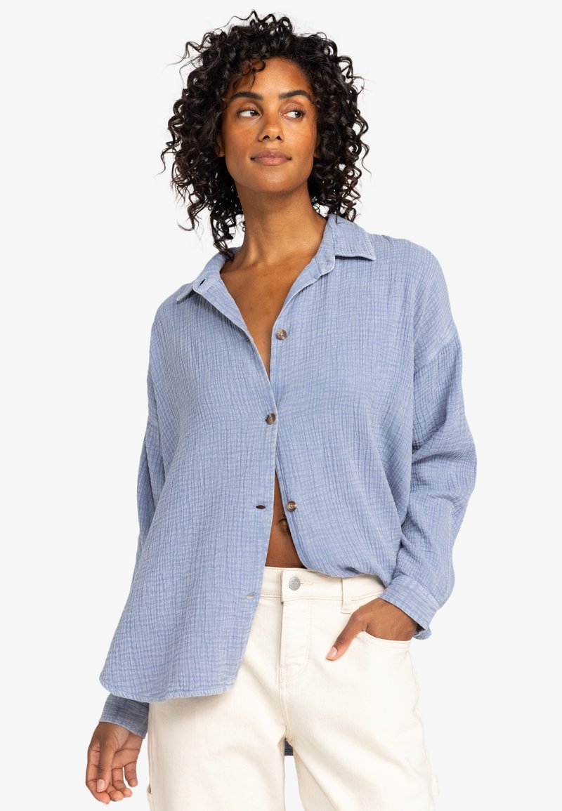 Light blue, textured button-up shirt with a loose fit, long sleeves, and a collar. Features brown buttons and an open front.