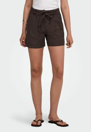 Kvinde iført mørkebrune shorts med høj talje og bundet bælte, hvid top, guldbøjler og sorte flip-flop sandaler, stående med benene let spredt.