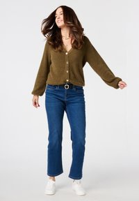 Olijfgroen fluffy cardigan met v-hals en knoopsluiting, gecombineerd met blauwe rechtgesneden jeans en witte sneakers. Gouden sieraden accenten zichtbaar.