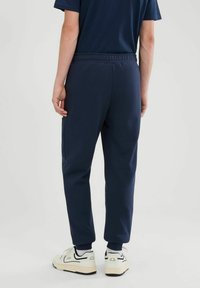 Osoba w granatowych dresowych spodniach, ciemnoniebieskiej koszulce, białych skarpetkach i białych trampkach stoi na jednolitym jasnym tle.