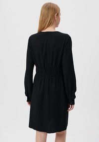 Schwarzes Kleid mit langen Ärmeln, geraffter Taille, strukturiertem Stoff und geknöpften Manschetten. Einfarbig, mittellanges Design, minimalistischer Stil.