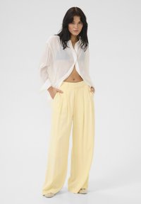 Camicia bianca trasparente con bottoni abbinata a pantaloni larghi giallo pallido, caratterizzati da pieghe frontali e tasche laterali, creando un look leggero e arioso.