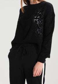 Zwarte gebreide trui met gestructureerde patronen en een losse pasvorm, aangevuld met decoratieve accenten aan de voorkant. Gecombineerd met zwarte broek.