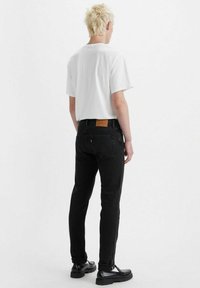 Jeans de mezclilla negra con un corte slim, parche trasero de cuero marrón claro y una camiseta blanca. El modelo está de pie con el cabello corto, mirando ligeramente hacia un lado.