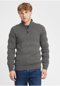 Suéter de color gris oscuro con cuello de media cremallera; presenta rayas horizontales texturizadas y puños y dobladillo acanalados. Se lleva con vaqueros azules.