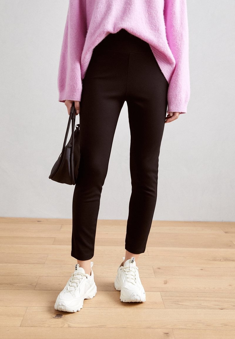 Black fitted pants with a smooth texture, paired with a pink knit sweater and white chunky sneakers, held with a small black bag.