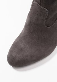 Botín de ante gris a la altura del tobillo con punta afilada y silueta curvada. Presenta detalles de costura y una textura suave y mate.
