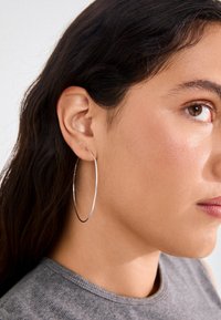Sølvørehængere, tynde og cirkulære, der viser en reflekterende metallisk finish. Båret på en models øre, med blødt, naturligt hår.