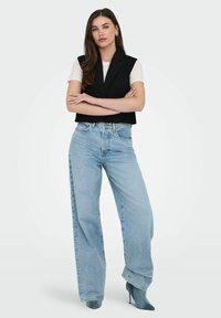Melna apgriezta veste virs baltas T-krekli, apvienota ar gaiši ziliem augstiem platām biksēm un norādītām tirkīza krāsas apaviem. Gluda auduma, ikdienišķs izskats.