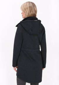 Šviesiaplaukė moteris vilkinti tamsiai mėlyną tekstūruotą striukę su gaubtu, matoma iš nugaros, stovinti prieš paprastą baltą foną.