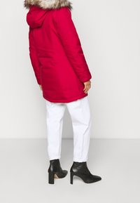 Czerwona parka z kapturem obszytym futrem, zapinana na zamek, z długimi rękawami i bocznymi kieszeniami, zestawiona z białymi spodniami i czarnymi botkami na obcasie.