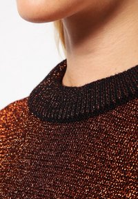 Suéter metálico bronce con un acabado texturizado, que presenta un cuello negro acanalado. Detalle de sutil brillo corto en toda la tela.
