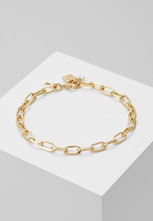 Bracelet chaîne en or avec des maillons rectangulaires, un petit pendentif perle et une étiquette carrée, exposé sur un socle blanc.
