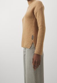 Beige Strickpullover mit hohem Kragen, gerippter Textur, seitlichen Schlitzen und Logo-Tag an der linken Naht. Kombiniert mit hellgrauen, weiten Jeans.