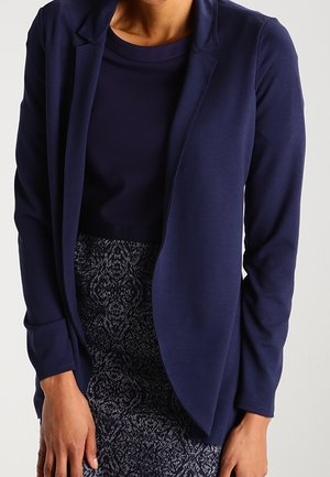 Frau trägt ein marineblaues Sakko über einem dunkelblauen Oberteil und einem hoch taillierten Rock mit einem schwarz-weißen, komplexen Muster.