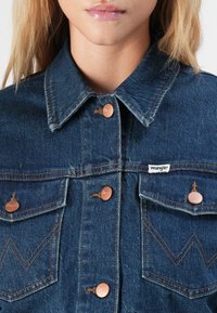 Närbild av person som bär en mörkblå denimjacka från Wrangler med kopparfärgade knappar och veckade fickdetaljer.