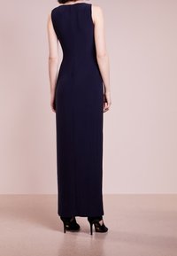 Navy sleeveless maxi dress with a fitted silhouette, smooth texture, and subtle pleats at the back, paired with black high-heeled shoes.