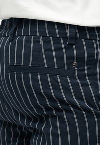 Gros plan sur un pantalon bleu foncé à fines rayures blanches verticales, une poche arrière, des passants de ceinture et un petit logo métallique près de la poche.