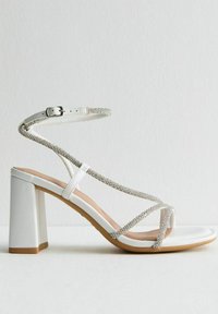 White high-heeled sandal with a sturdy block heel, featuring crossed straps adorned with silver embellishments and a buckle closure.