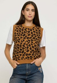 Femme avec de longs cheveux bruns portant un pull sans manches à imprimé léopard marron sur un t-shirt blanc et un jean bleu, les mains dans les poches.