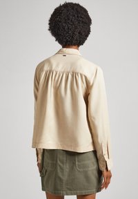 Cămașă bej cu mânecă lungă, cu spate plisat, manșete butonate și un mic logo pe guler. Asortată cu o fustă verde cu buzunare.