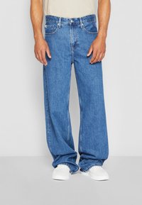 Zilas džinsu bikses ar platu kāju dizainu, ar pogu un rāvējslēdzēju, piecām kabatām un šuvju detaļām. Nēsātas ar baltām apaviem.