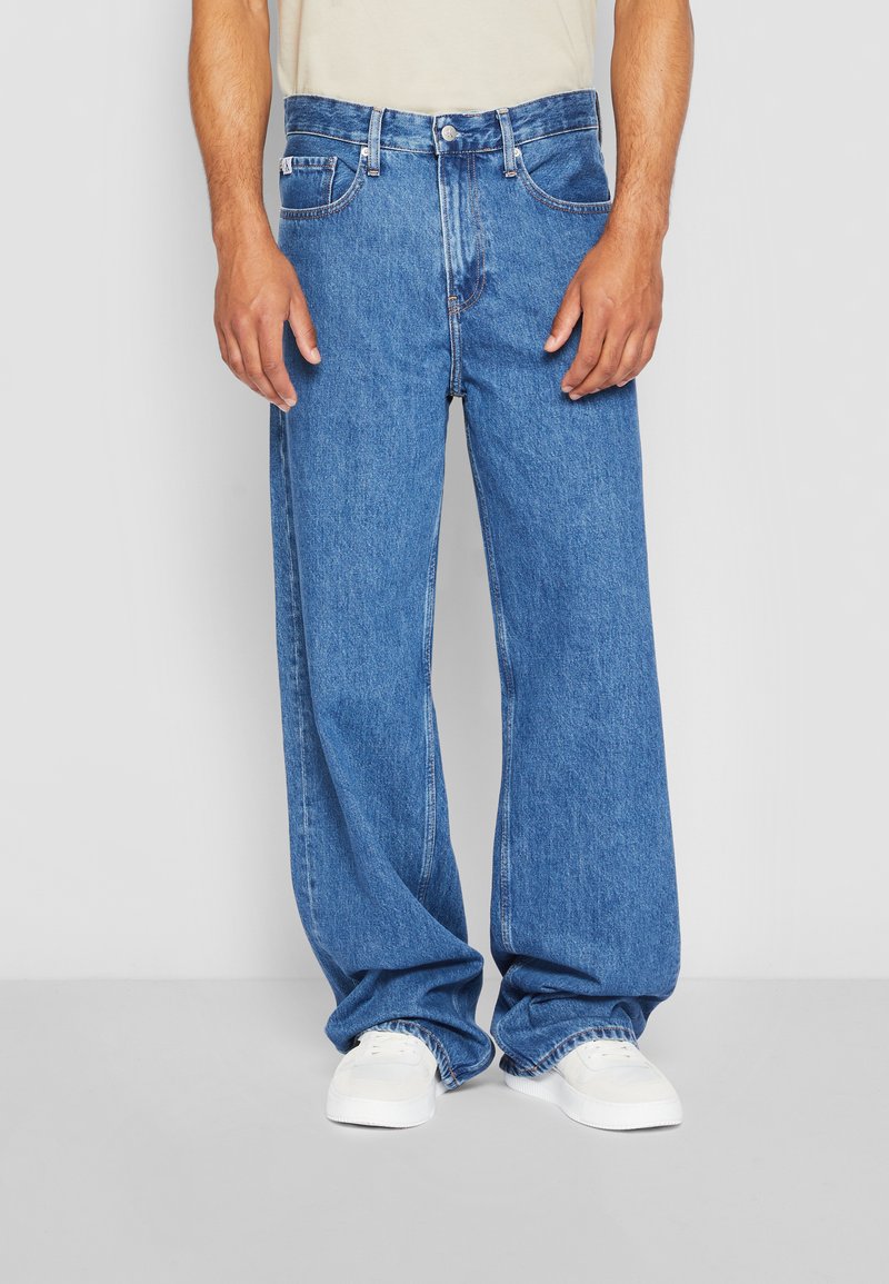 Zilas džinsu bikses ar platu kāju dizainu, ar pogu un rāvējslēdzēju, piecām kabatām un šuvju detaļām. Nēsātas ar baltām apaviem.