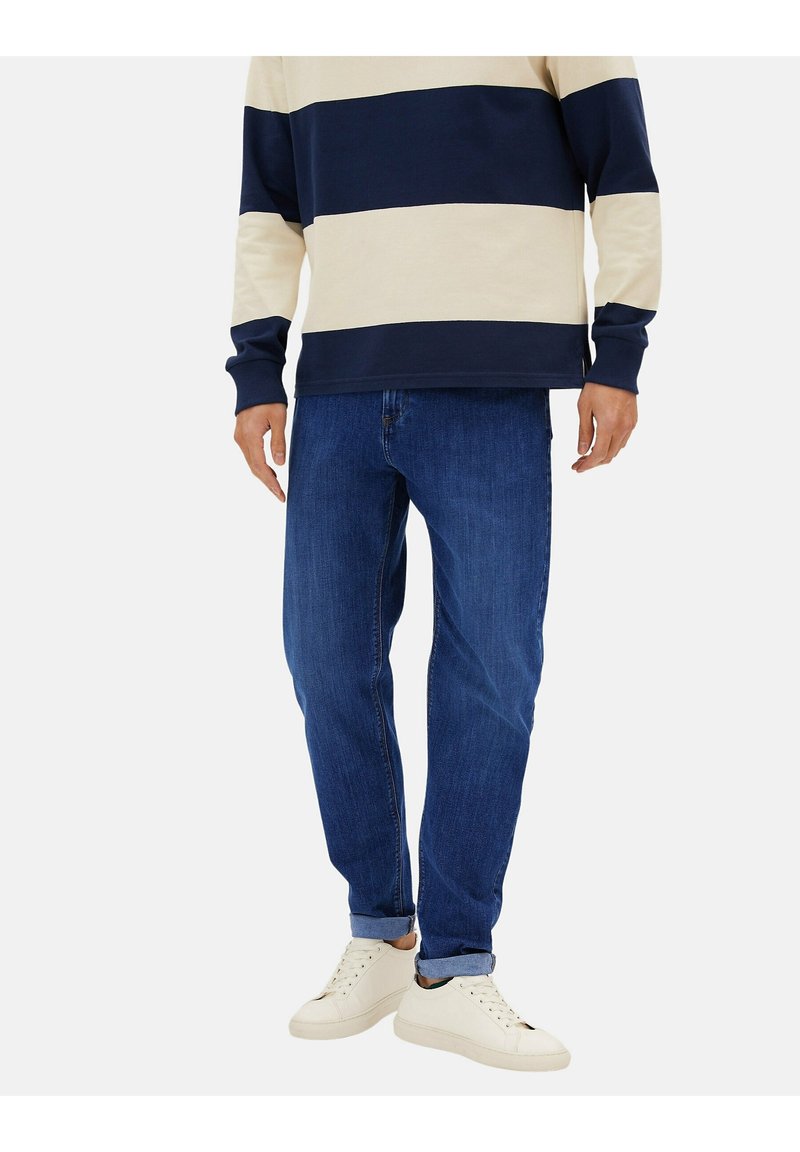 Jean en denim bleu à coupe droite, associé à un t-shirt à manches longues rayé crème et marine, et des baskets blanches. Texture douce, design décontracté.
