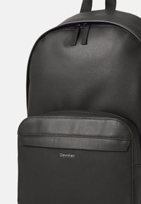Zaino nero realizzato in ecopelle texturizzata con un pannello frontale liscio e chiusure con zip. Presenta un logo Calvin Klein sul davanti.
