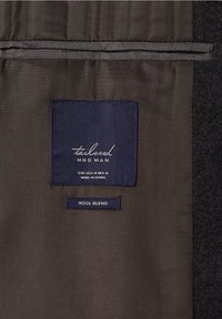 Wolmix stof in donkergrijs en bruin. Labels hebben blauwe accenten met tekst die het merk, de maat en de materiaalsamenstelling aangeeft.