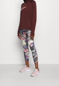 Burgunderfarbene Langarmoberteil mit einem weißen Nike-Logo, kombiniert mit bunten Leggings mit Blumenmuster und hellrosa Sportschuhen.