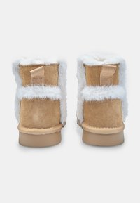 Botines de ante color beige con ribete de piel sintética blanca en la parte superior. Tienen forma redondeada, tacón plano y una pestaña trasera para facilitar el calce. Detalles de costura visibles.