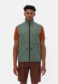 Gilet verde con zip frontale, colletto alto e due tasche anteriori, abbinato a una camicia color ruggine e pantaloni corti marroni. Tessuto leggero.