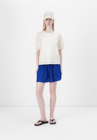 Vit topp med spetsmönster och puffärmar, i kombination med en blå broderad kjol och svarta flip-flops. Inkluderar även en matchande keps.