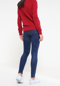 Rode sweatshirt met lange mouwen en geribbelde manchetten, gecombineerd met blauwe skinny jeans en witte sneakers, gezien vanuit de achterkant.
