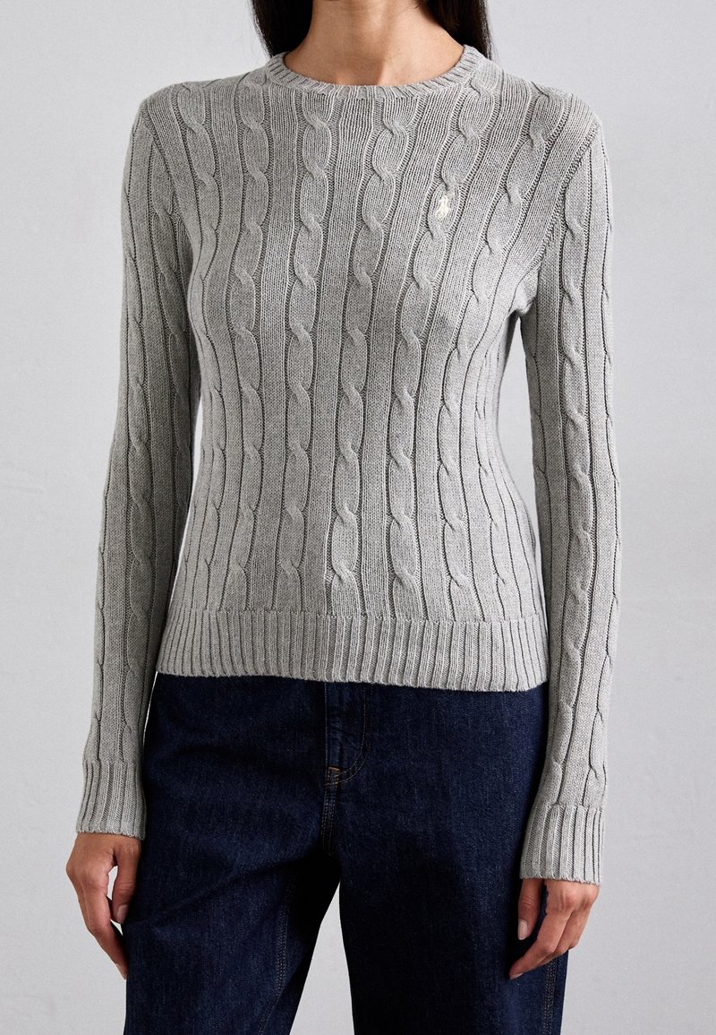 Femme portant un pull gris clair à torsades avec manches longues et un jean bleu foncé, posant devant un fond clair uni.