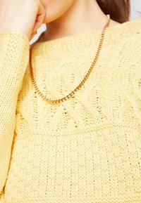 Žlutý pletený svetr s copánkovým vzorem, s texturovaným povrchem. Zlatý řetízkový náhrdelník zdobí výstřih.