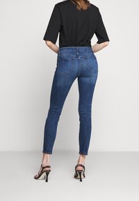 Mujer vistiendo jeans ajustados azul oscuro, camiseta de manga corta negra y sandalias de tacón abiertas negras, de pie contra un fondo claro.