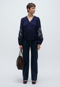 Μπλούζα ναυτικού μπλε με κεντητά λουλούδια στα μπαλόν μανίκια, συνδυασμένη με σκούρο τζιν καμπάνα και καφέ τσάντα ώμου με υφή.